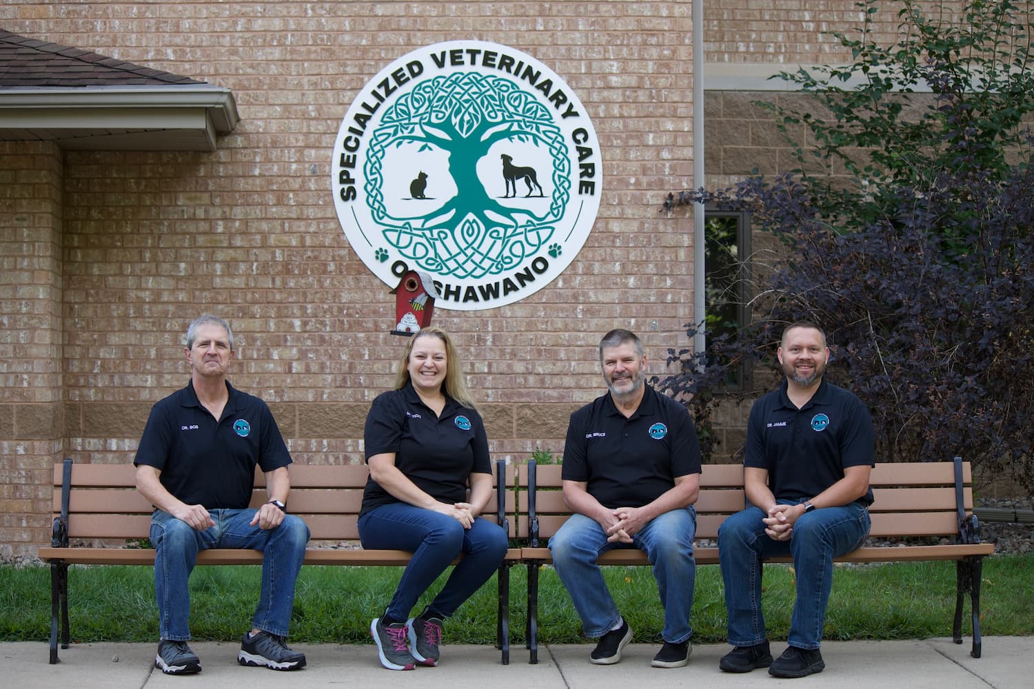 Four people wearing matching black shirts sit on benches in front of a brick building with a "Specialized Veterinary Care" sign.