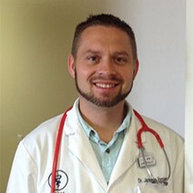 A person with short hair and a beard wearing a white coat and red stethoscope smiles at the camera.