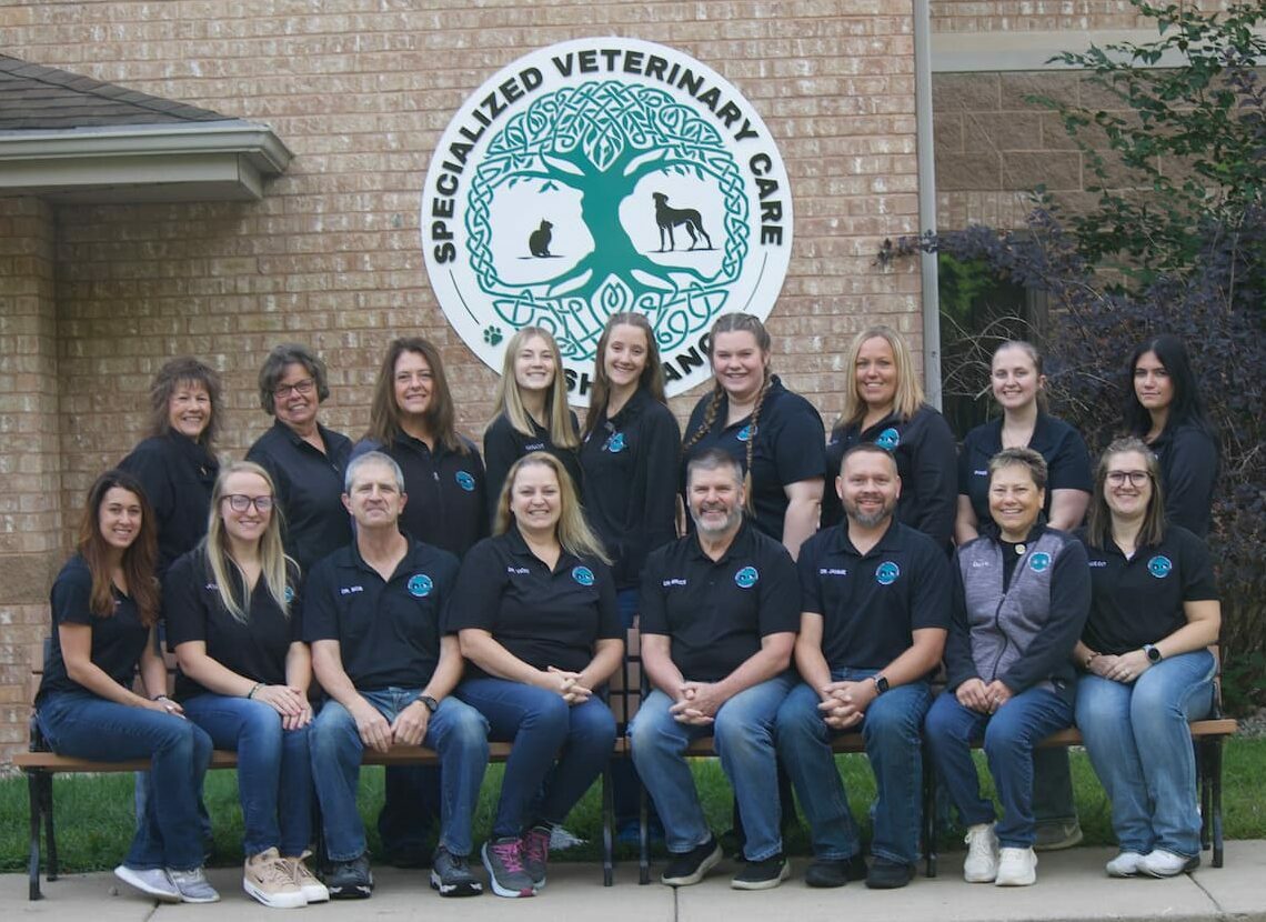 A group of sixteen people in matching black shirts sit and stand in front of a brick building with a "Specialized Veterinary Care" sign.