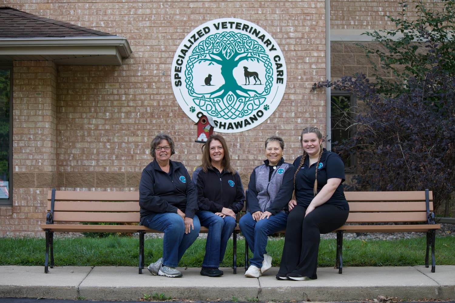 Four women sit on a bench in front of a veterinary clinic building with a sign that reads "Specialized Veterinary Care of Shawano.